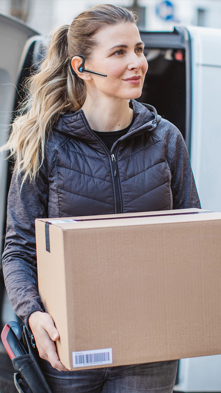 Delivery worker holding a large cardboard box in front of a van, wearing a dark quilted vest and a Bluetooth headset.