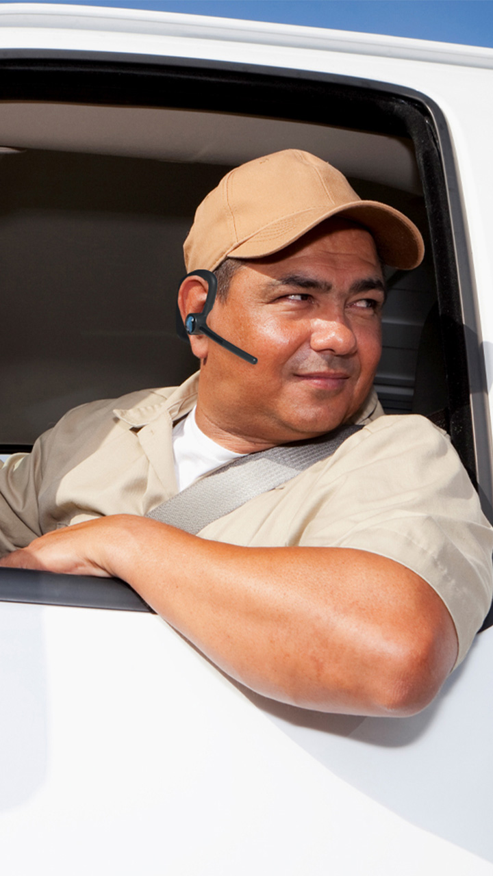 Man wearing the BlueParrott M500-XT Bluetooth and a beige baseball cap and shirt, seated in the driver’s seat of a white vehicle with a seatbelt on, left arm resting on the open window, wearing a wireless earpiece; face blurred.