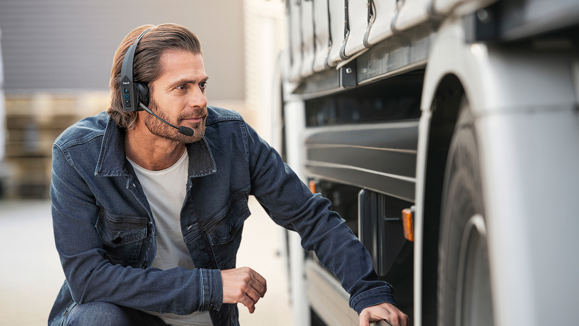 Person wearing a professional over‑ear BlueParrott Bluetooth headset kneeling beside a truck, inspecting the vehicle’s wheel area in an outdoor industrial setting.
